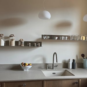 May include: A kitchen scene featuring two horizontal stainless steel wall shelves holding glass storage jars and glassware. Below, a light-colored countertop includes a sink, a faucet, a white fruit bowl with apples, and two tall shakers.
