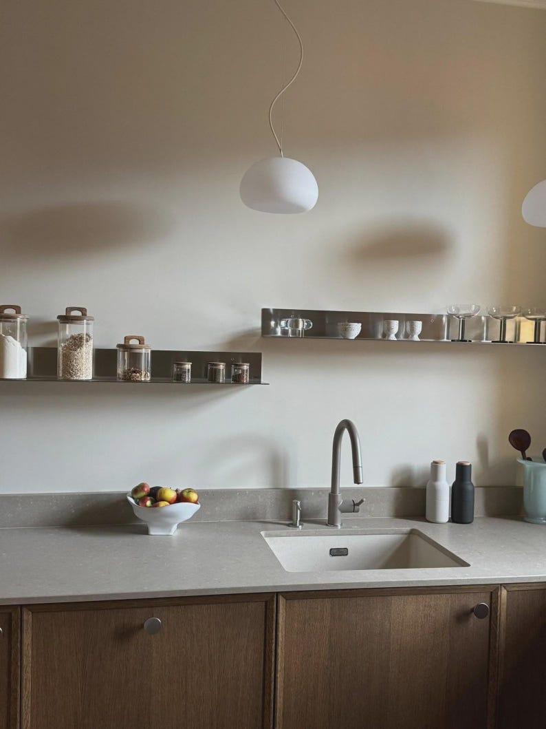 May include: A kitchen scene featuring two horizontal stainless steel wall shelves holding glass storage jars and glassware. Below, a light-colored countertop includes a sink, a faucet, a white fruit bowl with apples, and two tall shakers.