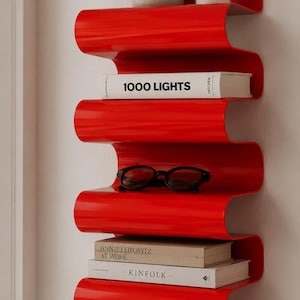 Könnte beinhalten: Eine leuchtend rote, wellenförmige Wandablage mit mehreren Ebenen, auf der Bücher, Brillen und Dekorationsartikel stehen. Auf einem Buch steht der Text "1000 LIGHTS". Eine stilvolle Aufbewahrungslösung.