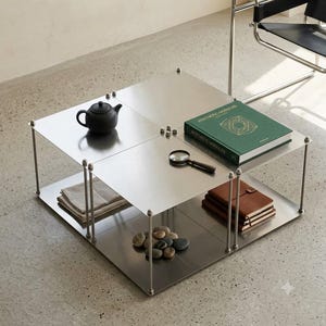 Può includere: Un tavolino quadrato a due livelli in acciaio inossidabile spazzolato con sottili aste di supporto verticali su ogni angolo. Il tavolo ospita una teiera nera, un libro verde intitolato Aesthetic Modules, una lente d'ingrandimento, tessuto piegato e pietre.