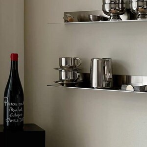May include: A dark wine bottle with a red cap and white text reading Parajes del Movastrell Ecologico Jumilla DO, positioned near two silver metal wall shelves holding stacked silver bowls, cups, saucers, a silver pitcher, and small items.