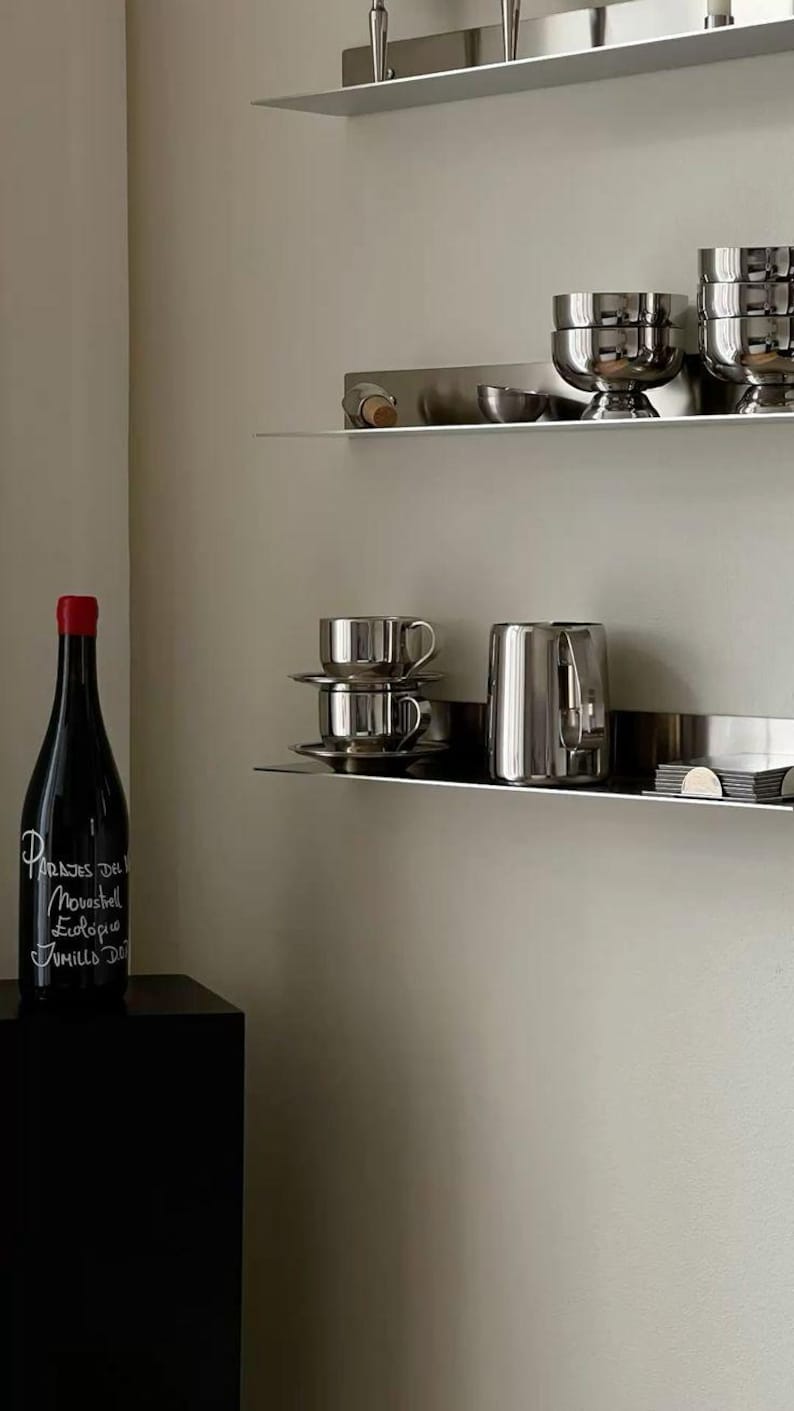 May include: A dark wine bottle with a red cap and white text reading Parajes del Movastrell Ecologico Jumilla DO, positioned near two silver metal wall shelves holding stacked silver bowls, cups, saucers, a silver pitcher, and small items.