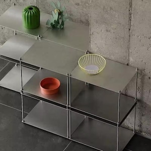 May include: A modular metal shelving unit with multiple rectangular shelves supported by thin vertical rods. The shelves hold a green ribbed vase, a green abstract sculpture, a yellow wire bowl, and a small orange bowl on the flat surfaces.