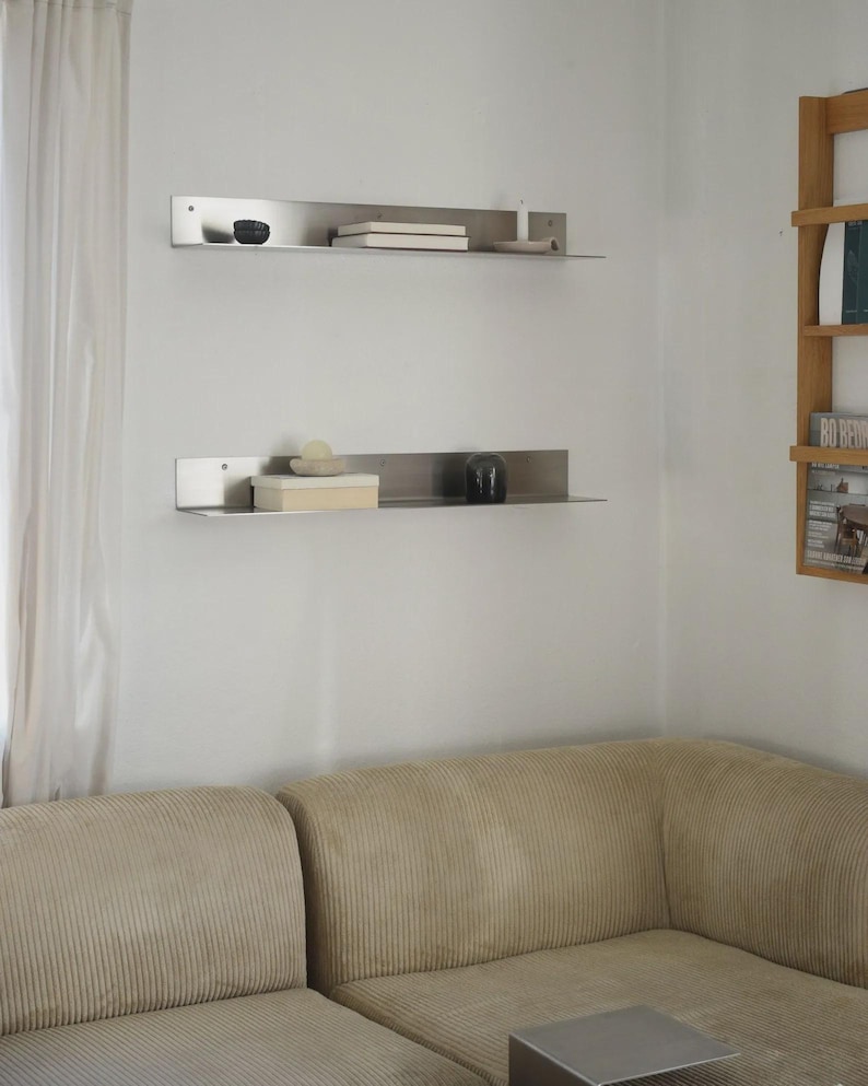 May include: Two long, horizontal, brushed stainless steel wall shelves mounted one above the other. Each shelf features a flat surface with a vertical back panel and visible screw holes for mounting, holding small decorative objects and books.