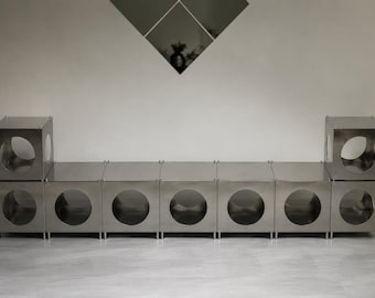 May include: A modular storage unit composed of nine metallic silver cubes arranged in a long horizontal row with two additional cubes stacked on the ends. Each cube features a large circular cutout on the front face, creating a uniform geometric design.
