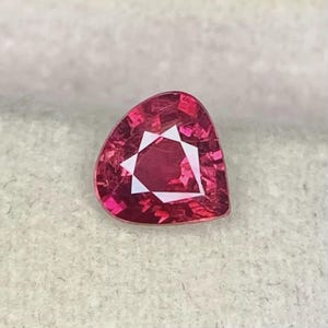 May include: A pear-shaped, faceted gemstone in a deep, vibrant red color. The stone features a complex internal geometric pattern with sharp, angular facets that reflect light, creating a brilliant, multi-faceted appearance across its surface.
