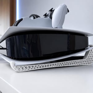 May include: A white 3D-printed stand with a hexagonal honeycomb pattern supporting a black and white gaming console. The console features a glossy black center panel flanked by white side plates, with a white wireless controller resting on top.