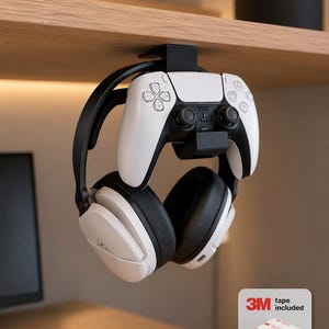 May include: A black under-desk mount holding a white and black gaming controller and a pair of white and black over-ear headphones. The mount is attached to the underside of a wooden shelf. An inset graphic shows a 3M adhesive strip with the text 3M tape included.