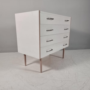Könnte beinhalten: Eine weiße Holzkommode mit vier horizontalen Schubladen und silberfarbenen Metallgriffen. Das Möbelstück steht auf vier konisch zulaufenden, hellen Holzbeinen und weist eine sichtbare Holzmaserung an den Schubladenfronten auf.