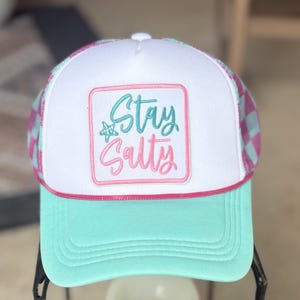 May include: A turquoise and white trucker hat with a pink and turquoise checkered pattern on the sides. The front panel is white with a square patch embroidered with the words "Stay Salty" in turquoise and pink.