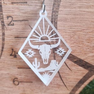 May include: A single, white, laser-cut earring with a diamond shape. The earring features a desert scene with a sun, bull skull, cactus, and geometric patterns. The earring hangs from a silver-colored hook.