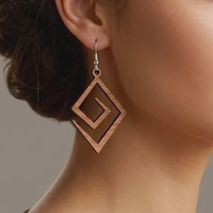 Peut inclure: Boucles d'oreilles géométriques en bois avec un motif en spirale carrée. Les boucles d'oreilles sont de couleur marron clair et sont suspendues à des crochets argentés. Elles sont portées par une personne aux cheveux foncés.