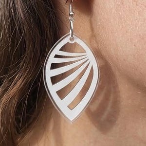 Peut inclure: Gros plan sur une boucle d'oreille argentée avec un pendentif blanc en forme de feuille. Le pendentif présente un motif découpé avec des lignes courbes rayonnant du centre. La boucle d'oreille est suspendue à un crochet argenté.