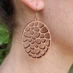 Peut inclure: Boucles d'oreilles ovales en bois clair avec un motif découpé de formes irrégulières. Les boucles d'oreilles sont de couleur marron clair et sont suspendues à un crochet argenté. Un accessoire unique.