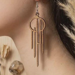 Peut inclure: Boucles d'oreilles géométriques en bois, composées d'un cercle et de lignes verticales pendantes. Les boucles d'oreilles sont de couleur marron clair et sont suspendues à un crochet argenté. Le fond comprend des plantes séchées, des pierres et de la lavande.