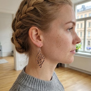 Peut inclure: Boucles d'oreilles pendantes en bois clair, en forme de feuille. Les boucles d'oreilles présentent une série de formes découpées. Elles sont suspendues à des crochets argentés. La personne porte un pull gris.