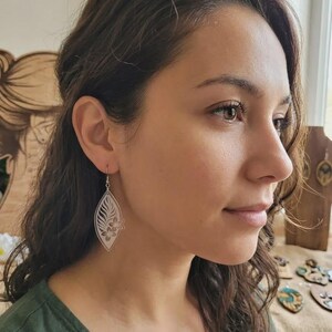 Peut inclure: Une femme porte des boucles d'oreilles argentées en forme de feuille. Les boucles d'oreilles présentent un motif floral découpé dans le métal. La femme porte un haut vert. L'arrière-plan est flou, mais semble être à l'intérieur.