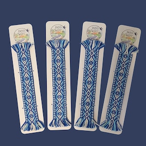 May include: Four woven bookmarks featuring a blue, light blue, and white geometric pattern. Each bookmark is attached to a white card with the text INTO THE WEFT and a small bird illustration at the top, finished with fringed ends.