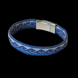 May include: A woven fabric bracelet featuring a repeating blue and white wave pattern. The band is secured with a polished silver-toned metal magnetic clasp. The design consists of interconnected geometric shapes in varying shades of blue.
