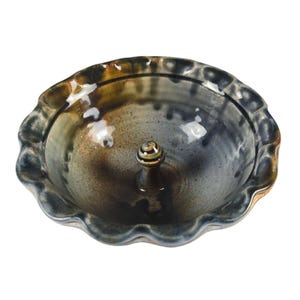 May include: A round ceramic ring dish with a scalloped edge and a central post. The glaze features a blend of dark blue, charcoal, and earthy brown tones with a glossy finish. The bowl has a deep basin and a wavy, textured rim design.