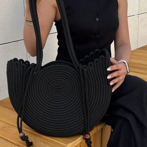 May include: A black handbag constructed from coiled rope, featuring a circular center design and a crescent-shaped silhouette. The bag includes two long shoulder straps and two dangling rope ends finished with small, dark wooden beads at the bottom.