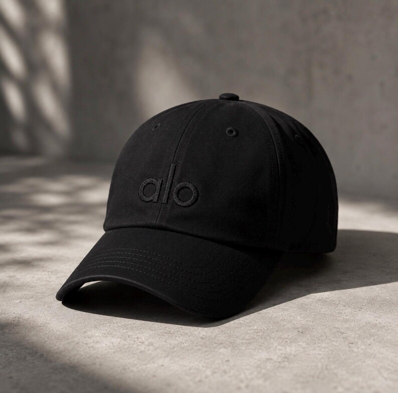 May include: A solid black baseball cap featuring a structured crown and a curved brim. The word alo is embroidered in matching black thread on the front center of the cap. The design includes a top button and stitched eyelets for ventilation.