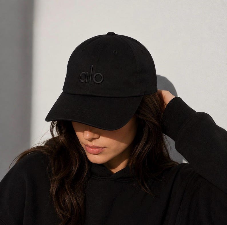 May include: A black baseball cap featuring the word alo embroidered in a matching black thread on the front panel. The cap has a structured crown, a curved brim, and a fabric strap at the back for adjustment, maintaining a monochromatic aesthetic.