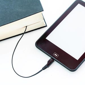 Può includere: Un e-reader nero con schermo bianco è collegato a un libro blu scuro tramite un cavo nero. L'e-reader è su una superficie bianca. Il libro è chiuso.