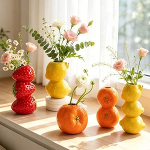 Puede incluir: Una colección de cinco jarrones de cerámica con forma de fruta que incluye una pila de fresas rojas, una pila de limones amarillos, una naranja individual, una esfera naranja y una pila de limones amarillos, con flores rosas y follaje.