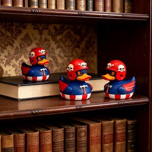 May include: Three rubber ducks with dark blue bodies, orange beaks, and red wings. Each duck wears a red helmet featuring white skull and crossbones symbols. The chest of each duck displays a small white hammer icon above red and white stripes.