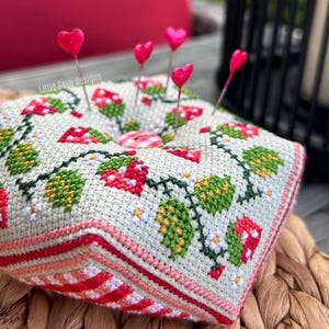 May include: A square pincushion with a cross-stitched design featuring red mushrooms, green leaves, and white flowers. Pink heart-shaped pins are inserted into the cushion. The pincushion has a red and white striped border. The text "Little Dove Designs" is visible.