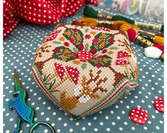 May include: A cross-stitched pincushion with a woodland design featuring mushrooms, trees, and a squirrel. The pincushion is beige with red, green, and brown accents. Red and white polka dot buttons and scissors are also visible.