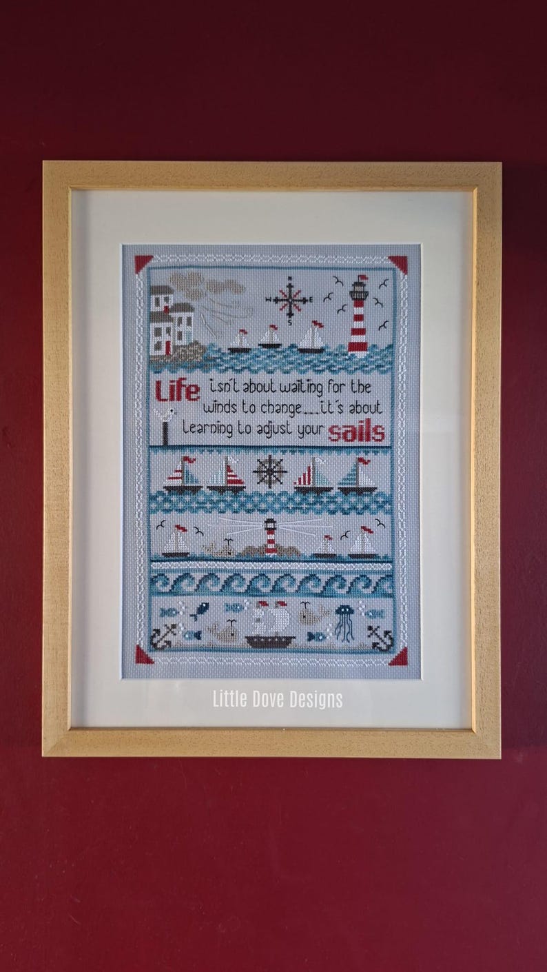 May include: Framed cross-stitch art with a nautical theme. The design features sailboats, a lighthouse, and the text "Life isn't about waiting for the winds to change...it's about Learning to adjust your sails." The frame is light brown.