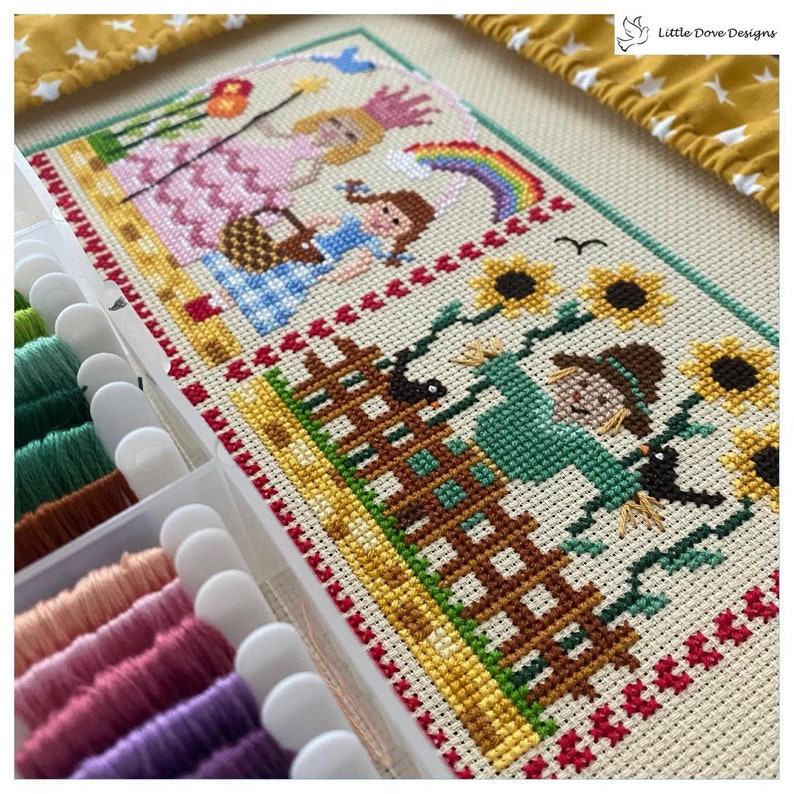 Puede incluir: Un proyecto de bordado de punto de cruz que presenta una escena caprichosa con una princesa, una ni&ntilde;a con un vestido azul, un arco&iacute;ris, un espantap&aacute;jaros y girasoles. El bordado est&aacute; parcialmente completado y se muestra sobre un fondo blanco con un contenedor de hilos de bordar.