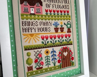 May include: A framed cross-stitch sampler on beige fabric featuring a pink house, flowers, a watering can, and a sun. The text reads A GARDEN FULL OF FLOWERS BRINGS MANY HAPPY HOURS. The design includes red, blue, yellow, and green thread colours.
