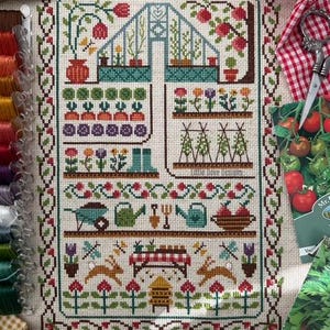 Peut inclure: Une broderie au point de croix sur tissu clair représentant une scène de jardin avec une serre, des rangées de légumes, des fleurs, une brouette, des arrosoirs, des lapins et une ruche, le tout entouré d'une bordure florale décorative.