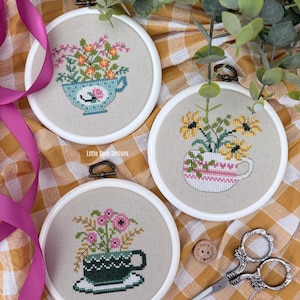 May include: Three cross-stitch embroidery hoops featuring floral designs in teacups. The teacups are in blue, pink, and green, each holding different flowers. The text "Little Dose Designs" is visible. A pair of decorative scissors and a wooden button are also present.