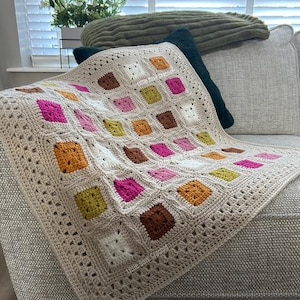 May include: A crocheted throw blanket with a cream background and colourful squares in shades of pink, orange, brown, and green. The blanket has a delicate crocheted border.