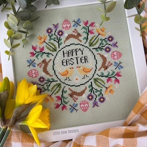 May include: A square framed cross-stitch embroidery on sage green fabric featuring the text HAPPY EASTER in black, surrounded by a circular wreath of colourful flowers, brown rabbits, patterned eggs, and two yellow birds, with Little Dove Designs text.