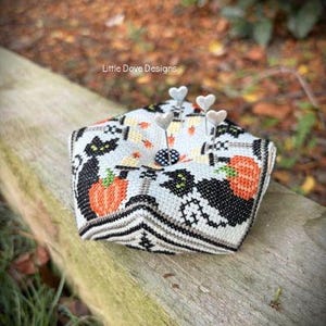 May include: A pentagonal pincushion featuring cross-stitch embroidery with black cats, orange pumpkins, and white candles on a light blue background. The centre has a black and white polka dot button with four silver heart-topped sewing pins.