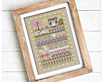 May include: A framed cross-stitch sampler featuring a spring theme with a house, sheep, ducks, birds, flowers, and umbrellas. The text reads "SPRING AWAKENING" and "MARCH APRIL MAY". The design is bordered by yellow flowers on a tan fabric background.