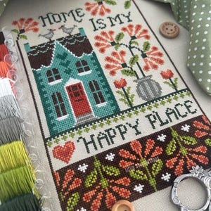 May include: Cross-stitch embroidery with a teal house, red door, and the text "HOME IS MY HAPPY PLACE." The design includes flowers, birds, and a heart. Wooden buttons and embroidery floss are also visible.