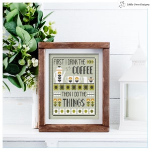 May include: A framed cross-stitch design with a green background and yellow flowers. The text reads "First I drink the coffee then I do the things".