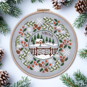 Peut inclure: Un cercle de broderie au point de croix représentant une scène hivernale avec une maison, des arbres et le mot "WINTER". Le motif est entouré d'une couronne de verdure, de baies et de pommes de pin. Le cercle est en bois et est posé sur un fond enneigé.