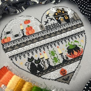 May include: A heart-shaped cross-stitch design with Halloween imagery. The design features a haunted house, pumpkins, spiders, black cats, and a bubbling cauldron. The colour palette includes orange, black, grey, and white. The design is surrounded by fabric and embroidery floss.