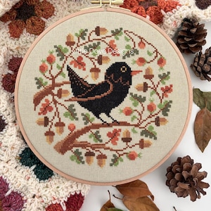 Op de afbeelding: Een ronde houten borduurring met een kruissteekontwerp van een zwarte vogel op een tak. De vogel is omringd door een krans van eikels, oranje bessen en groene bladeren. De tekst Little Dove Designs is zichtbaar.