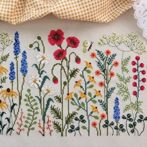 May include: A cross-stitch embroidery pattern on beige fabric featuring a variety of wildflowers including red poppies, blue spikes, white daisies, and yellow blossoms. A small wooden button and copper-colored stork scissors are placed nearby.