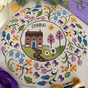 Puede incluir: Un diseño de punto de cruz con temática primaveral. El diseño presenta una casa, ovejas y un camino, rodeados por una corona floral. La palabra "SPRING" está en la parte superior. La paleta de colores incluye azules, amarillos, rosas y verdes. Hilo de bordar y cinta en primer plano.