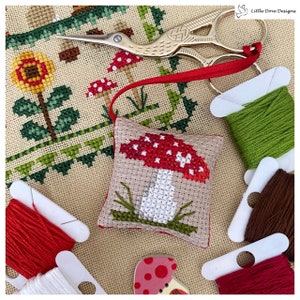 May include: A cross-stitched mushroom ornament with a red cap and white spots, resting on a tan fabric. Green and brown embroidery floss, a pair of gold scissors, and a red ribbon are also visible. The background features a cross-stitched floral design.