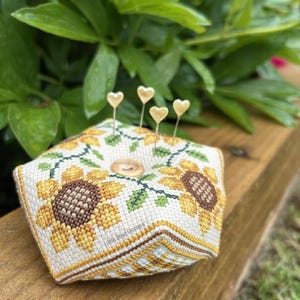 Puede incluir: Un cojín cuadrado bordado a punto de cruz con un diseño de girasoles. El cojín beige tiene girasoles amarillos y marrones, hojas verdes y alfileres dorados en forma de corazón. El texto "Little Bova Designs" es visible.